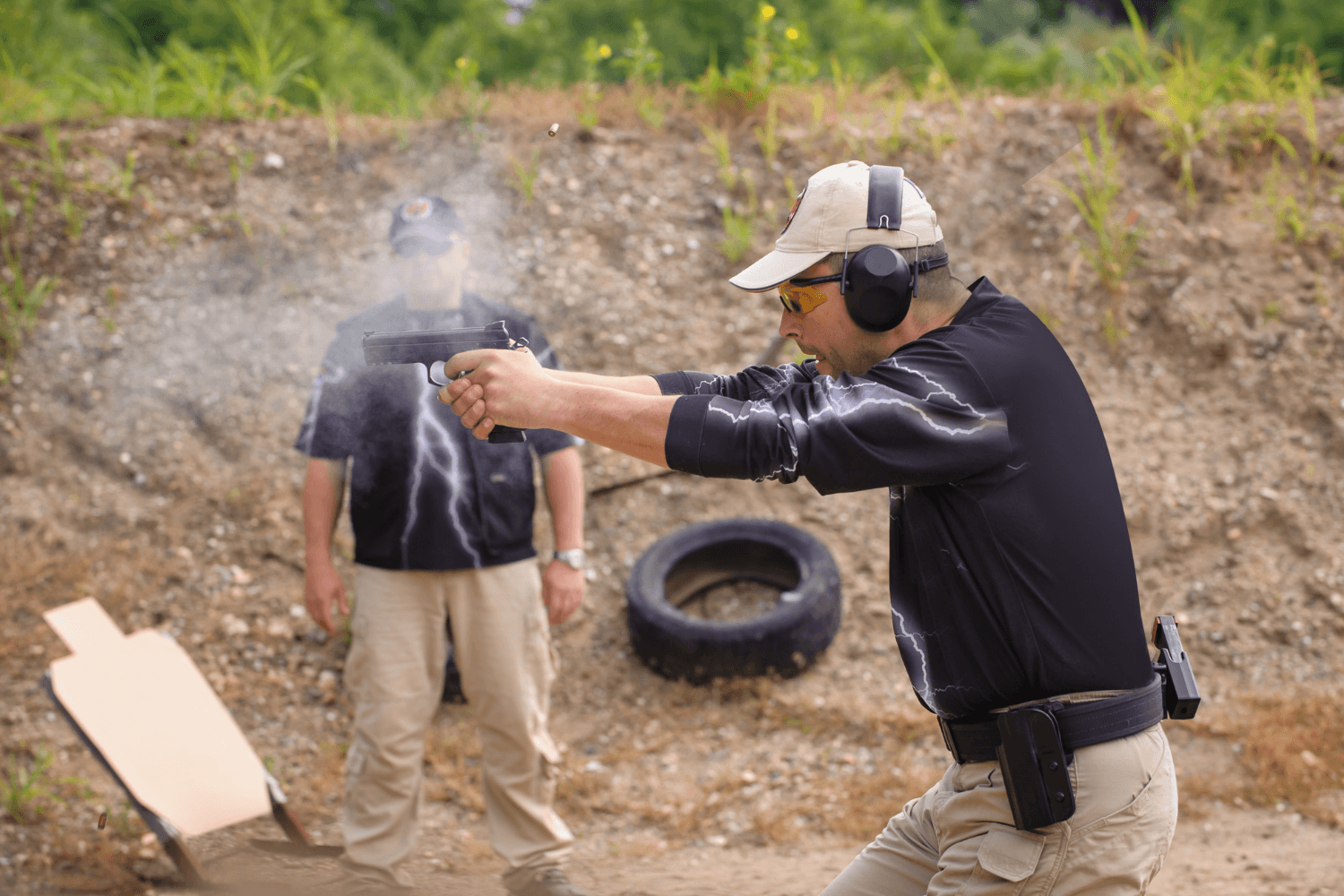 Man firing his pistol at an outdoor shooting range as his partner watches on